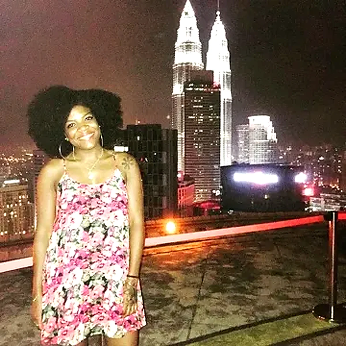 Woman in a floral dress on a rooftop with the Petronas Towers lit up behind her